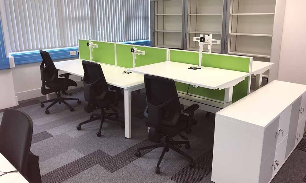 Modern office with white desks, green privacy panels, black chairs, and a row of windows along the left wall.