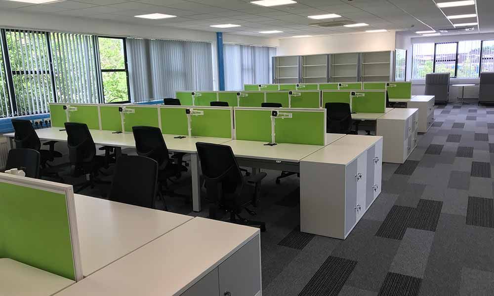 Empty office with rows of cubicles, green partitions, black chairs, and large windows.