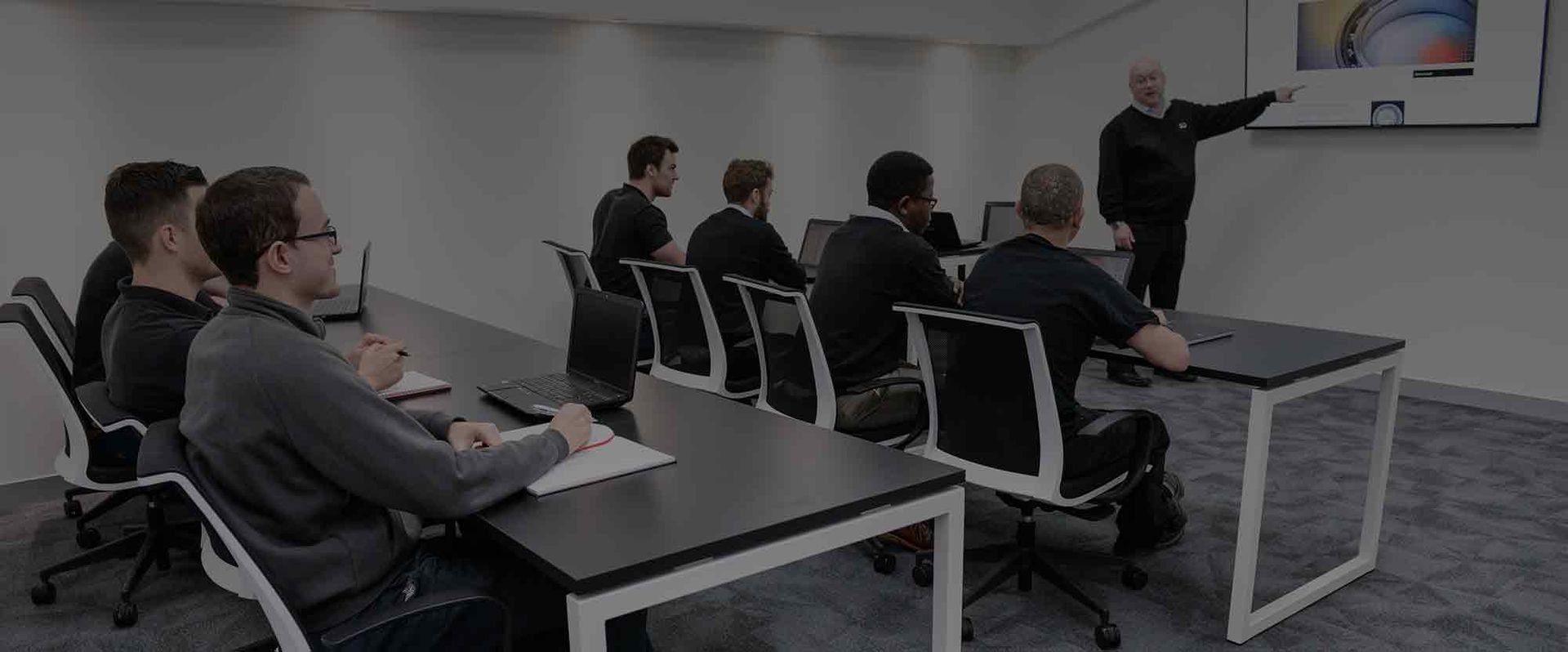 People seated in a classroom watch a presenter pointing at a projected screen.