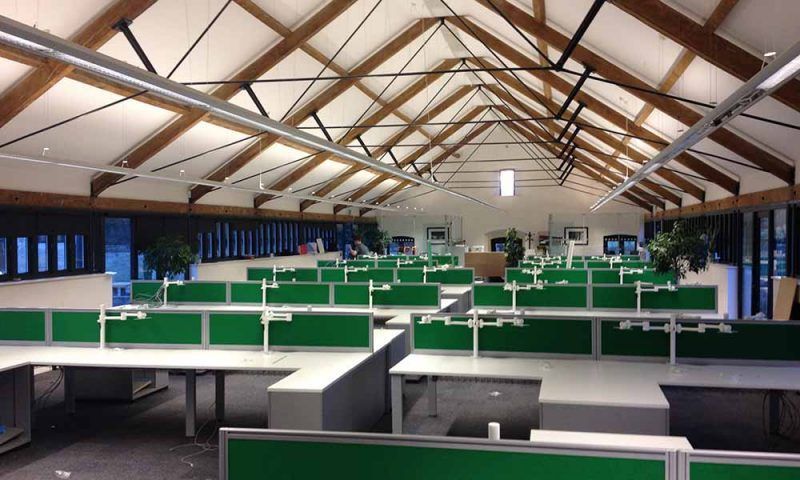 Empty office cubicles under a high wooden-beam ceiling with green partitions and white desks