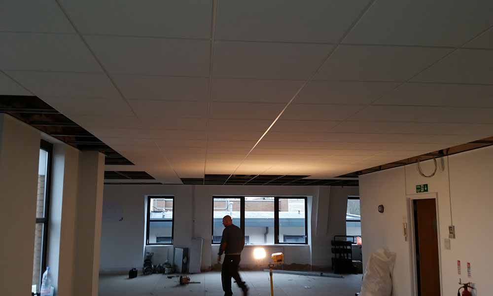 Empty office interior with a person standing under exposed ceiling lights near windows.