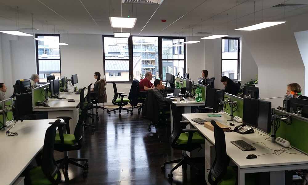 Open-plan office with people working at desks and computers near large windows.