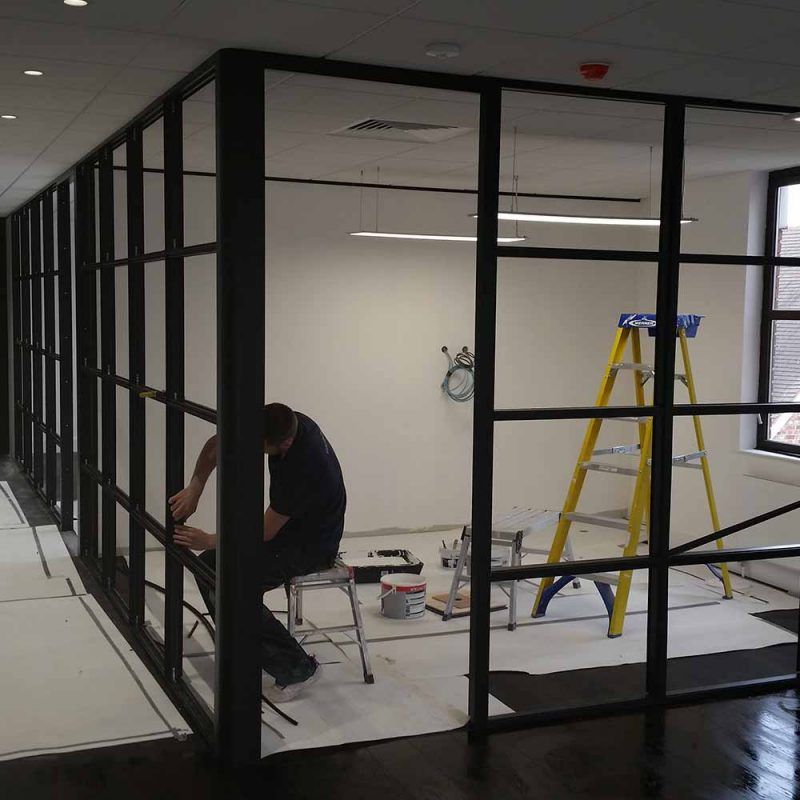 Worker installing a black-framed glass wall in an office under renovation, with a yellow ladder nearby.