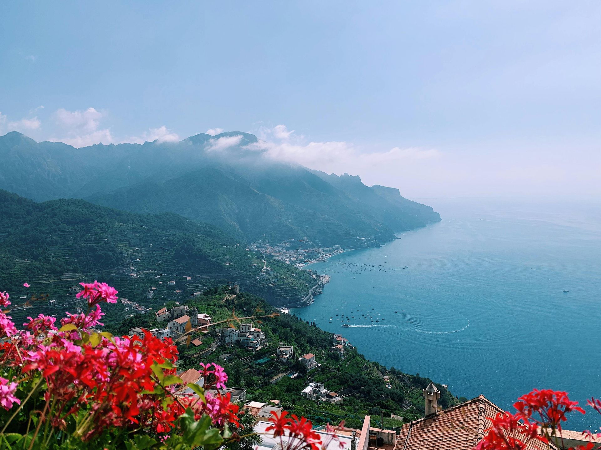 Coastal view with pink flowers, buildings on a hillside, and a calm, blue sea under a clear sky on the Amalfi Coast in Italy.