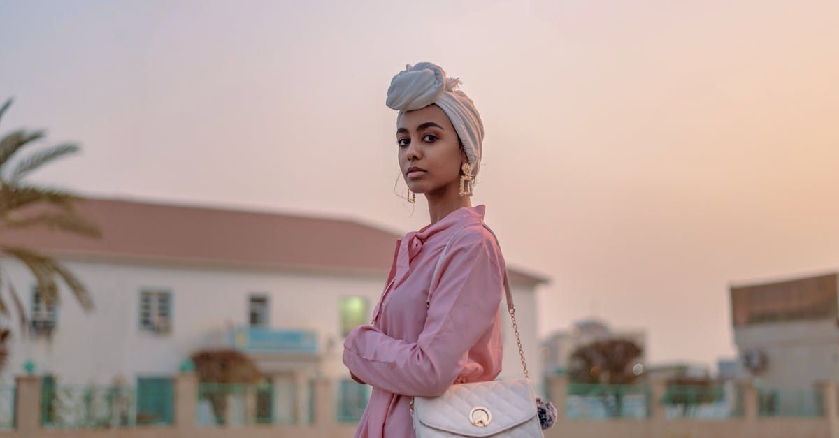 Woman wearing a head wrap and pink shirt, arms crossed, outside near a building with a palm tree, sunset in background.