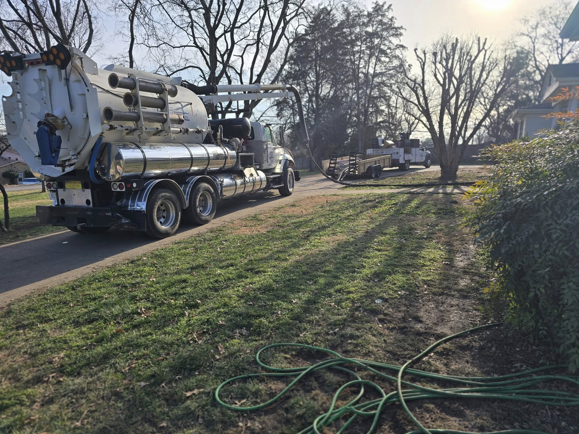 A large white hydro-excavation truck parked on a residential street near a grassy lawn and a garden hose.