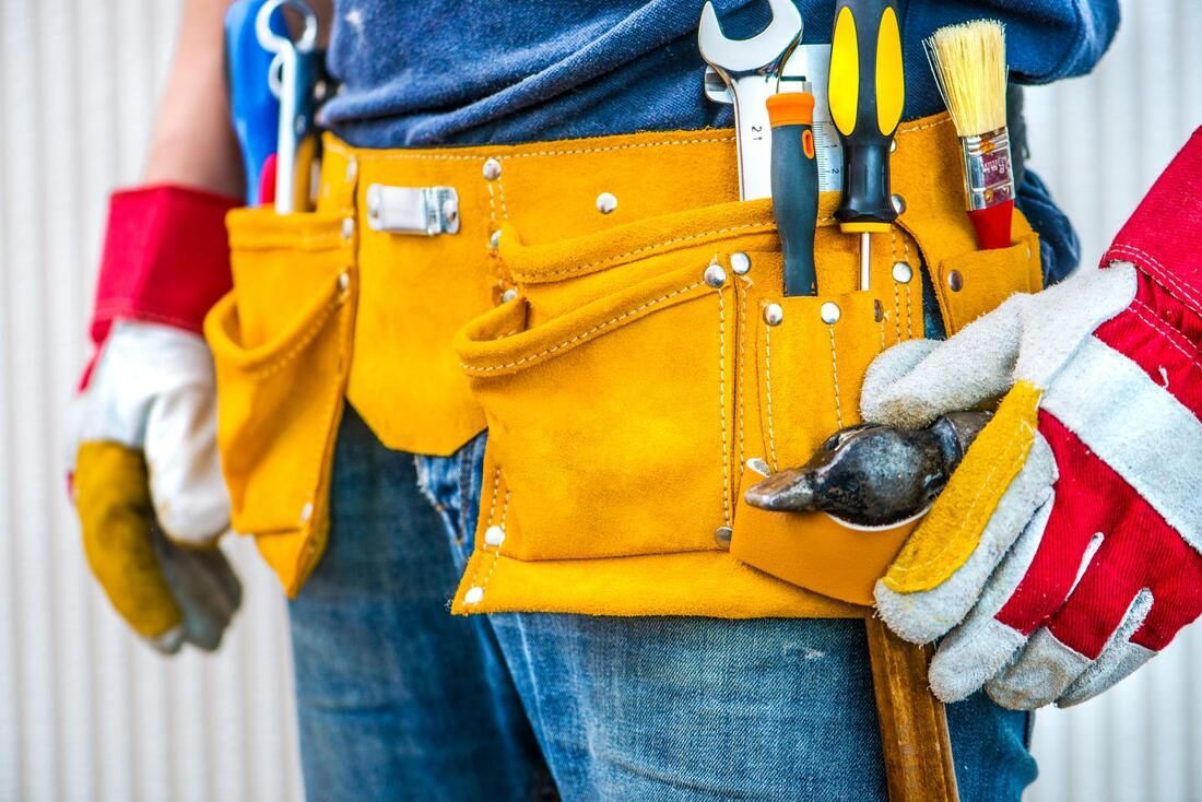 Worker wearing a yellow tool belt with tools and red-and-white work gloves, standing in jeans and a blue shirt
