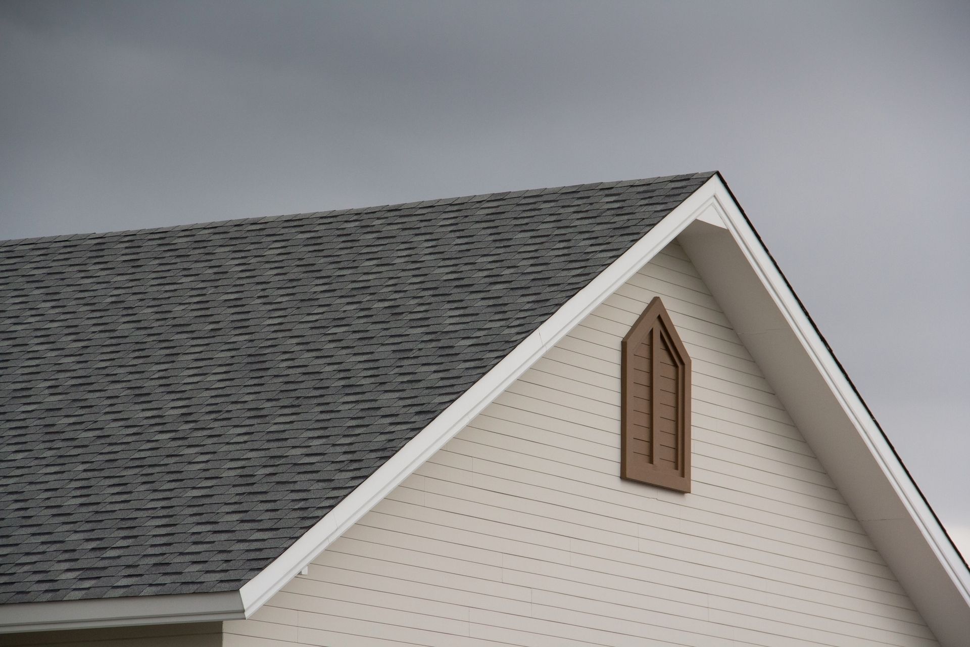 Gray-shingled roof and white siding of a building under a cloudy sky. Brown, arched window detail.