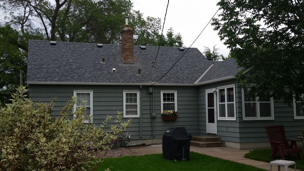 Green house with dark gray roof, chimney, white-framed windows, and a black grill in backyard.