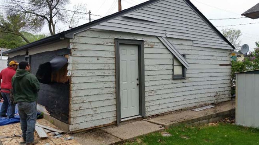 Men removing siding from a gray, weathered garage with a white door.