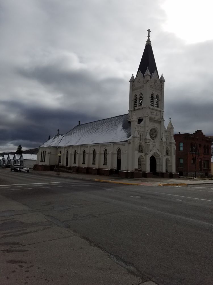 White church with tall spire under cloudy sky. Snow on roof, building on corner.