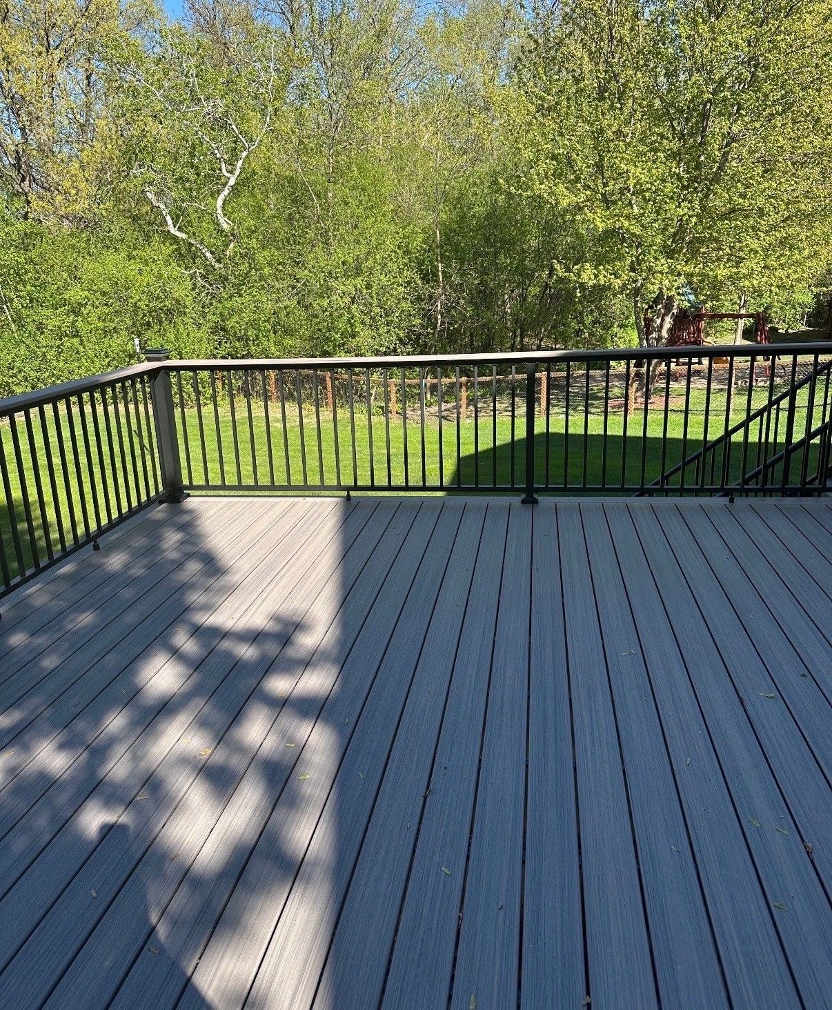 Wooden deck with black railing overlooking a green yard and trees in bright sunlight.