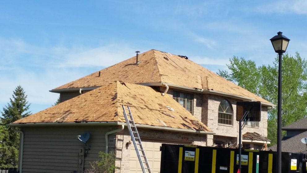 House with exposed roof under construction, ladder, and a dumpster on a sunny day.