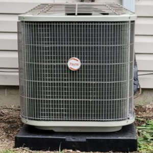 Outdoor air conditioner unit beside a beige vinyl wall with a gray electrical box above it