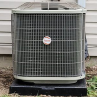 Outdoor air conditioner unit beside a beige vinyl wall with a gray electrical box above it