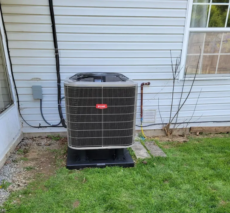 Outdoor HVAC condenser unit beside a house, set on a black pad near siding and grass