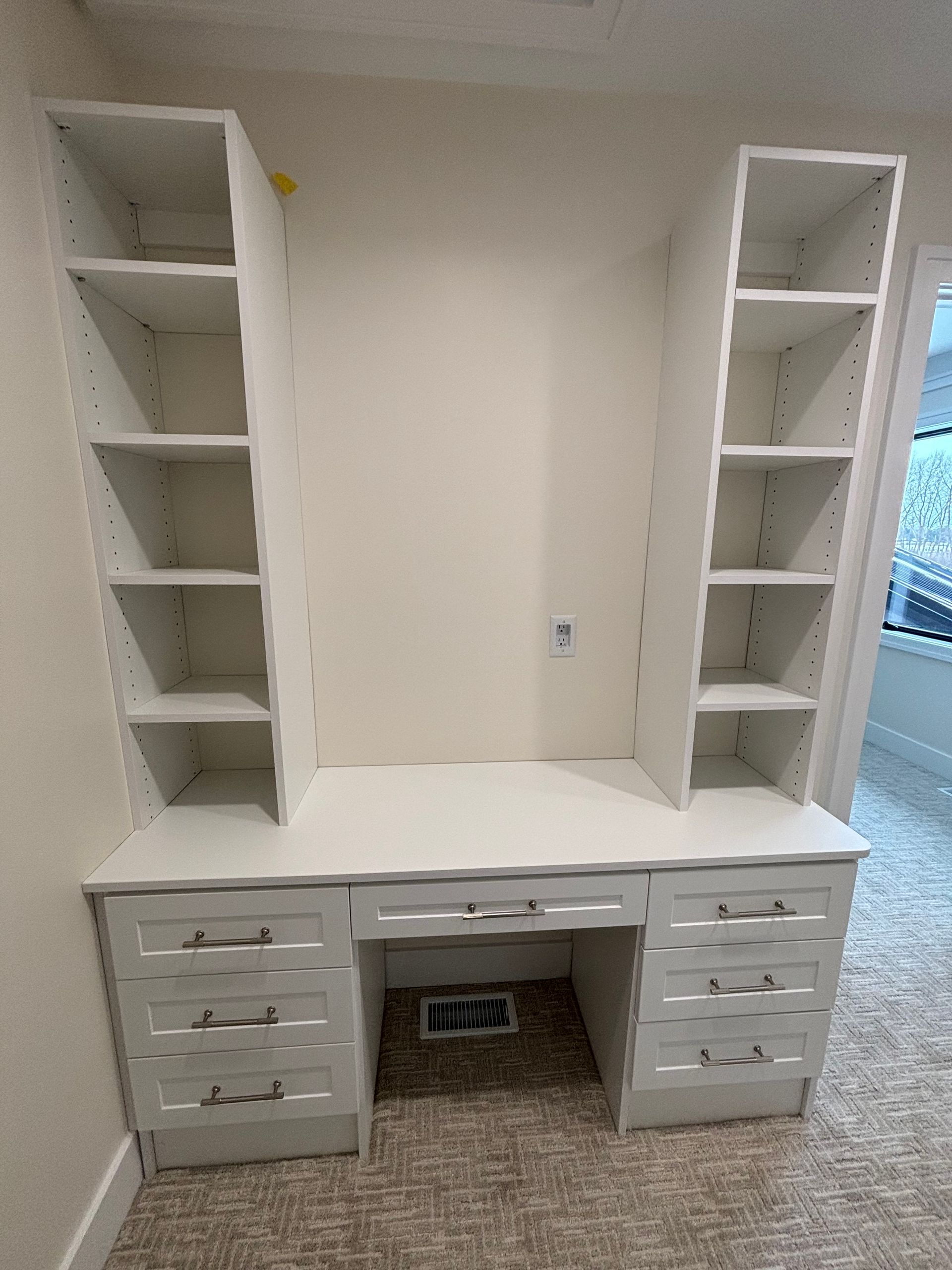 White built-in desk with drawers and tall shelving in a room, unfinished and near a window