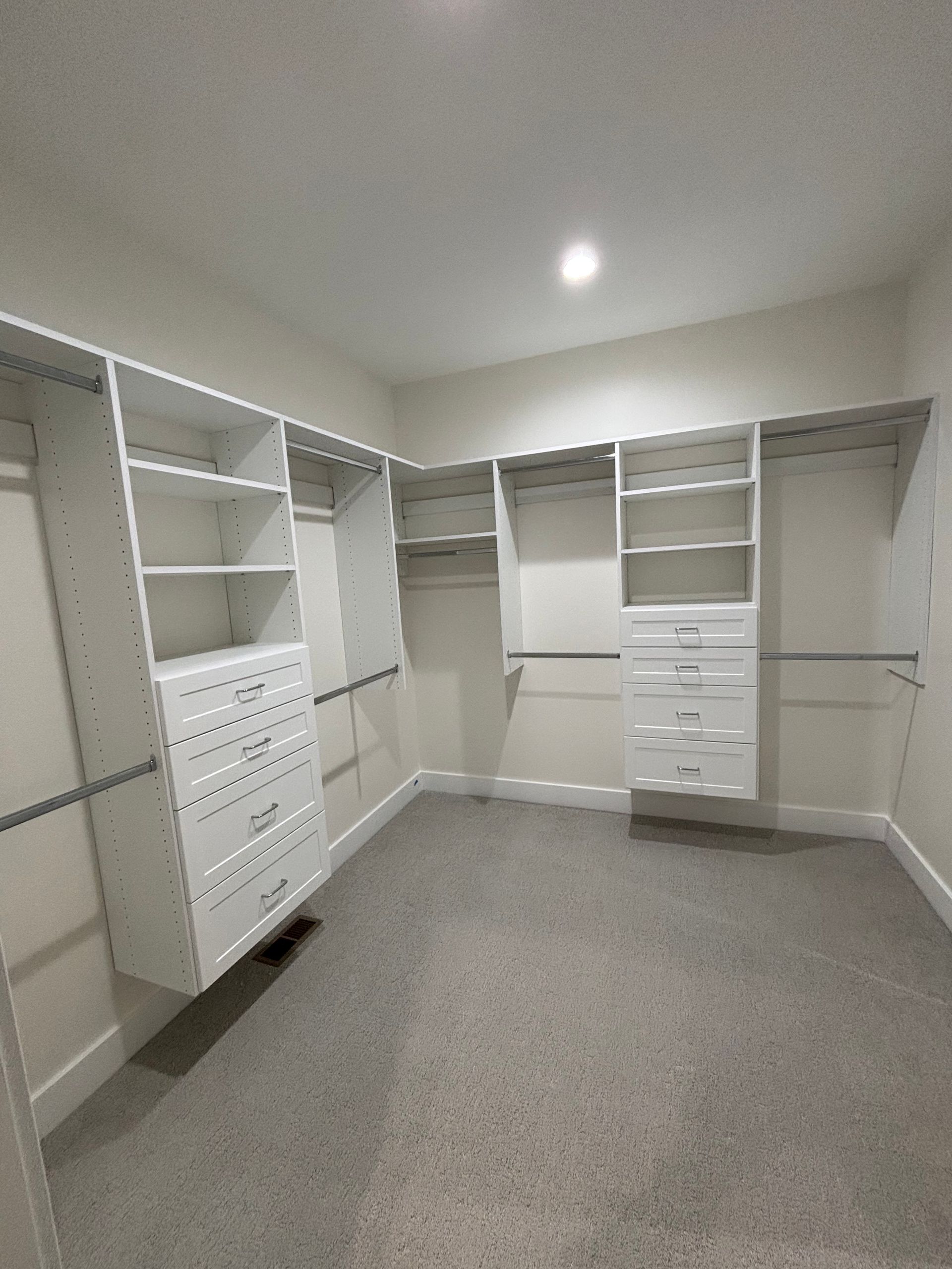 Empty walk-in closet with white built-in shelves and drawers, gray carpet, and a ceiling light.
