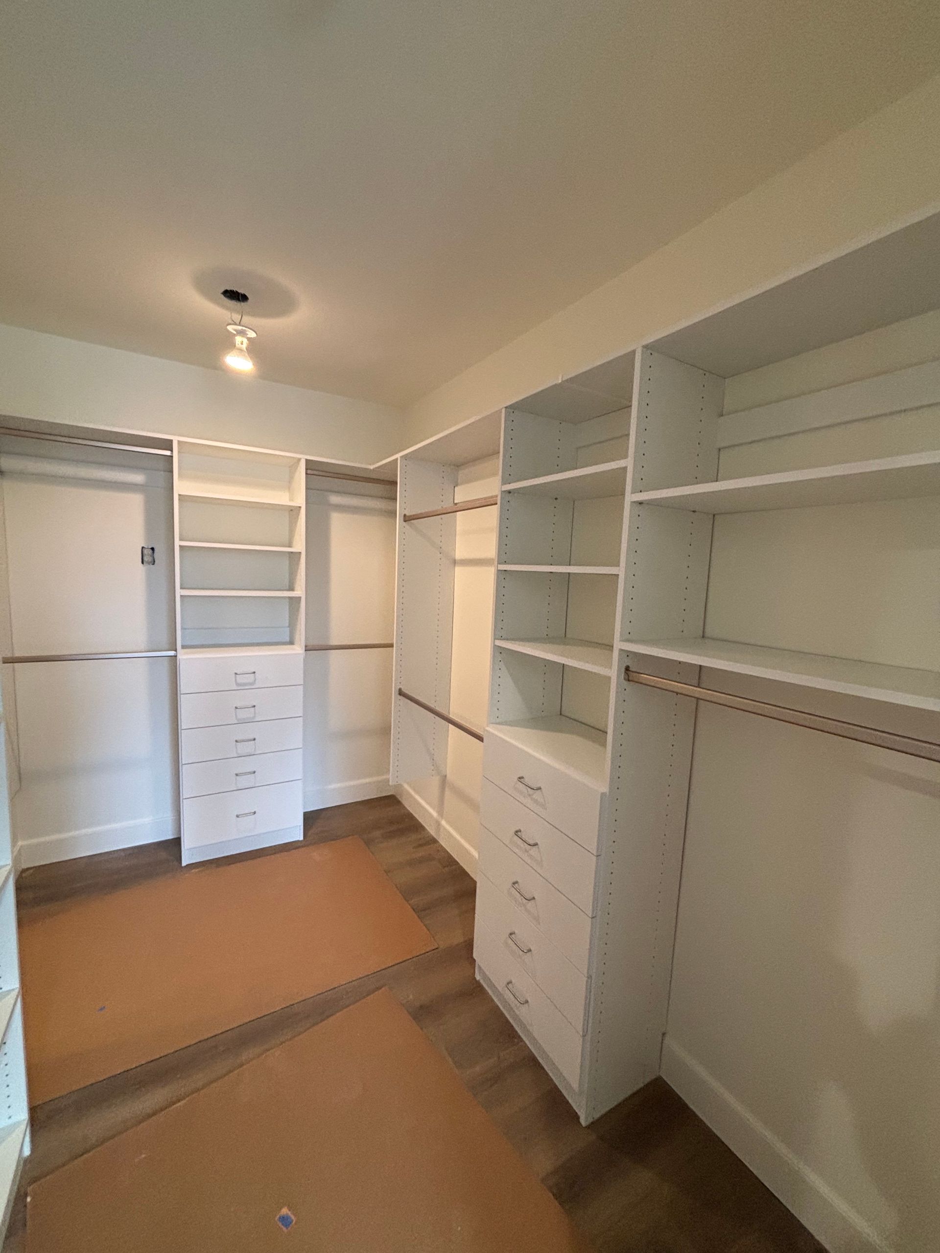 Empty walk-in closet with white shelves, drawers, and hanging rods, brown carpet, and a ceiling light.