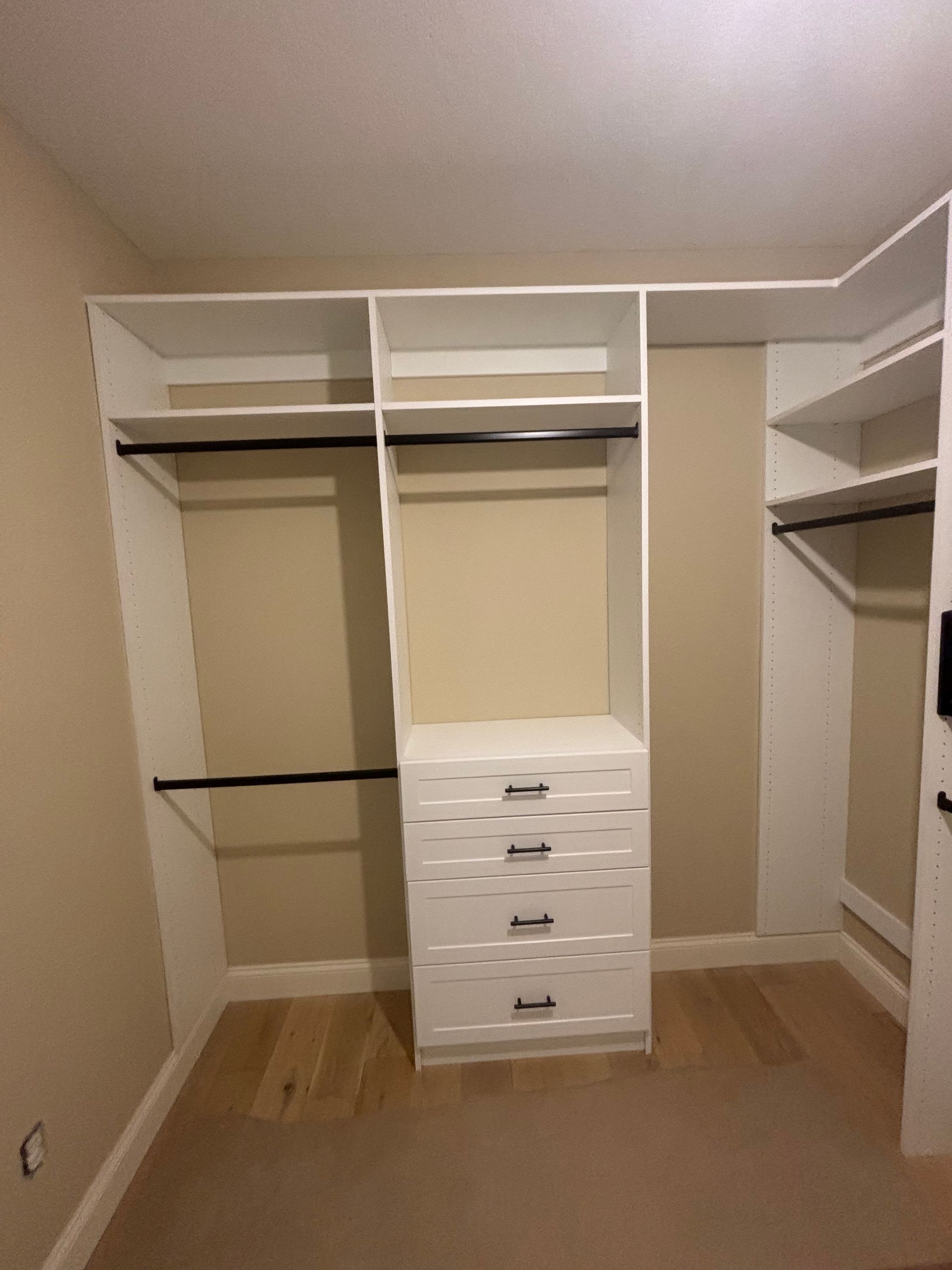 Empty walk-in closet with white shelving, hanging rods, and a center drawer chest on wood flooring