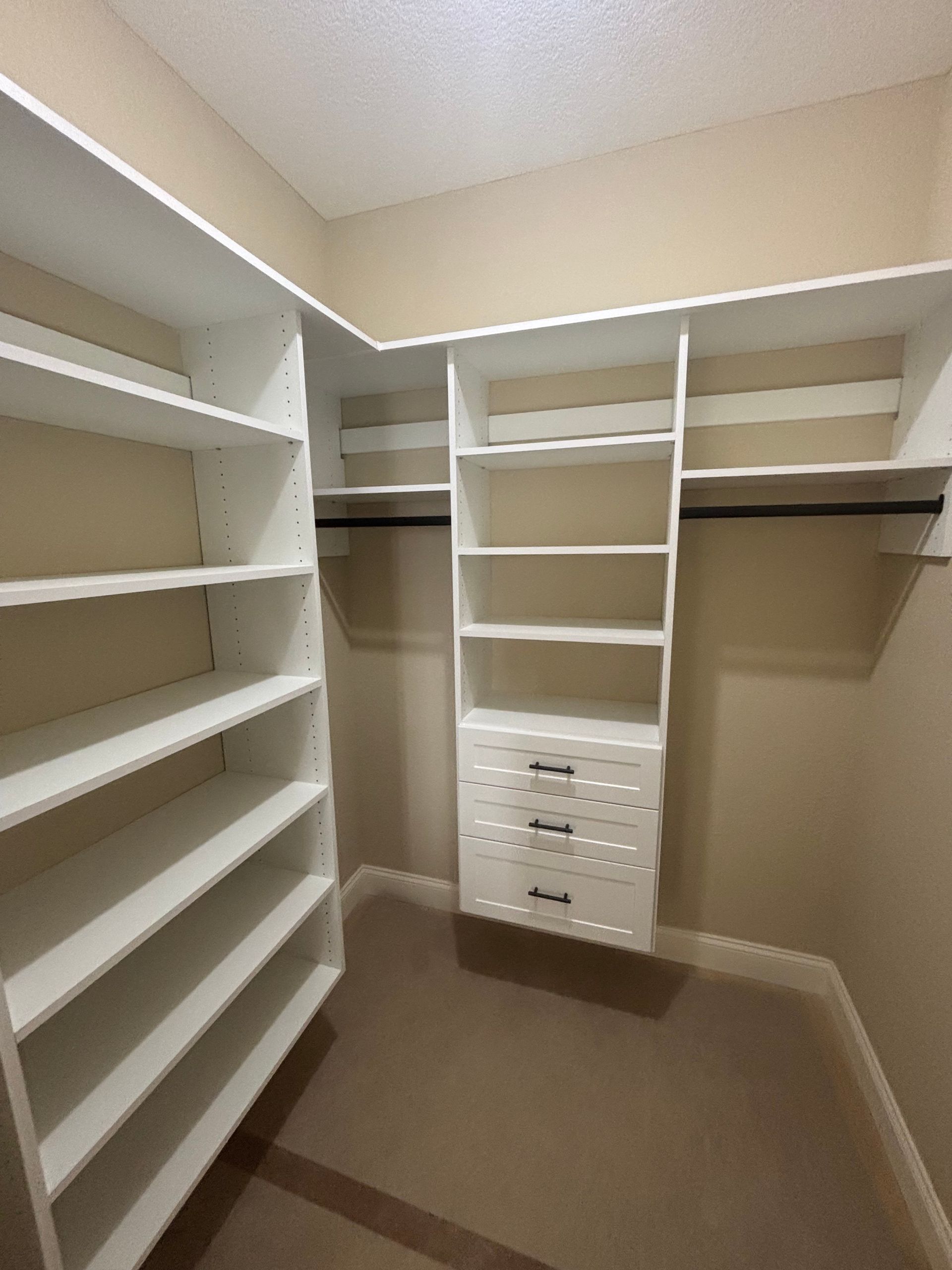 Walk-in closet with white shelving, hanging rods, and center drawers on beige walls and carpet