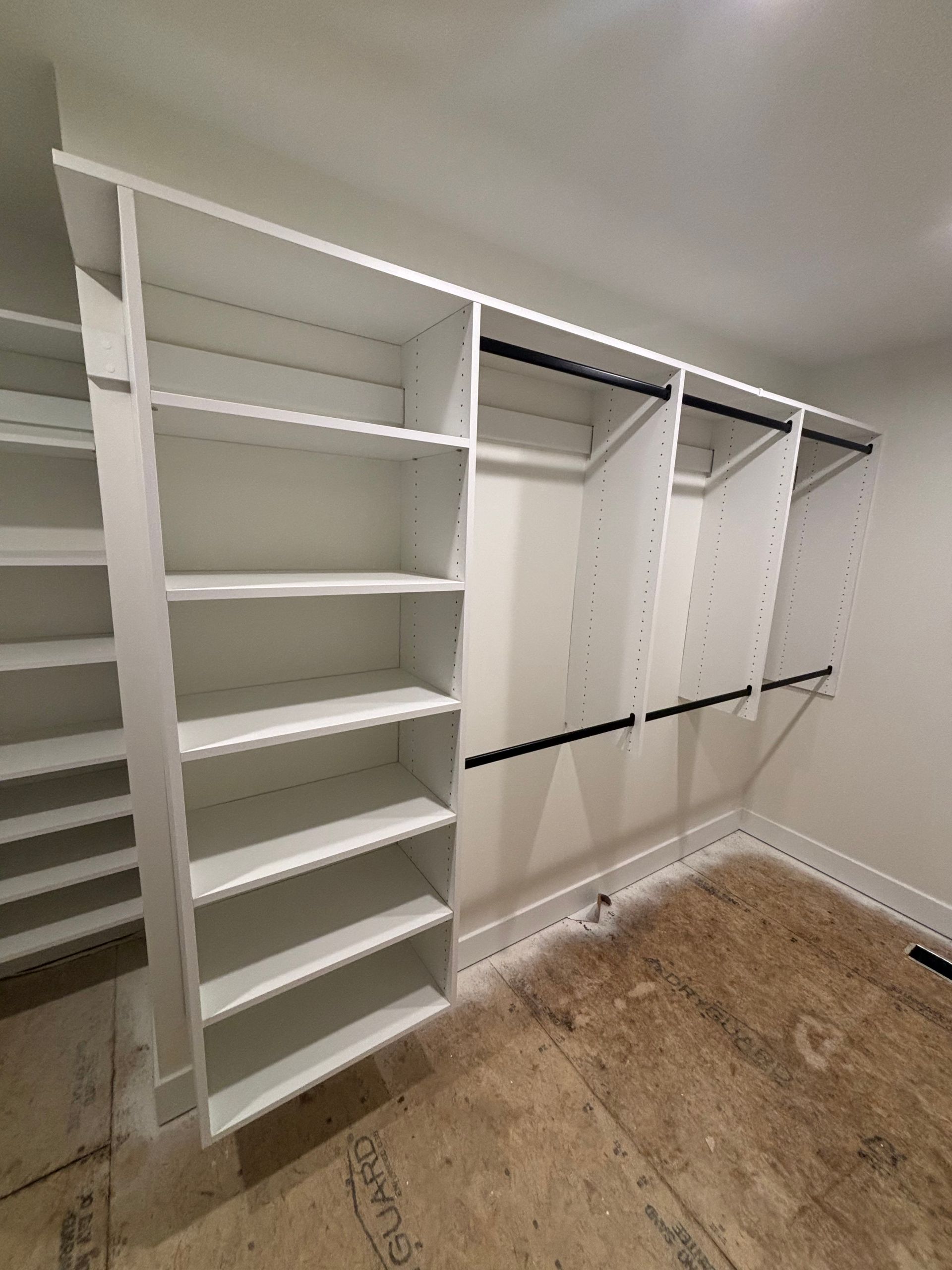 White built-in closet with shelves, hanging rods, and open storage in an empty room with unfinished floor.