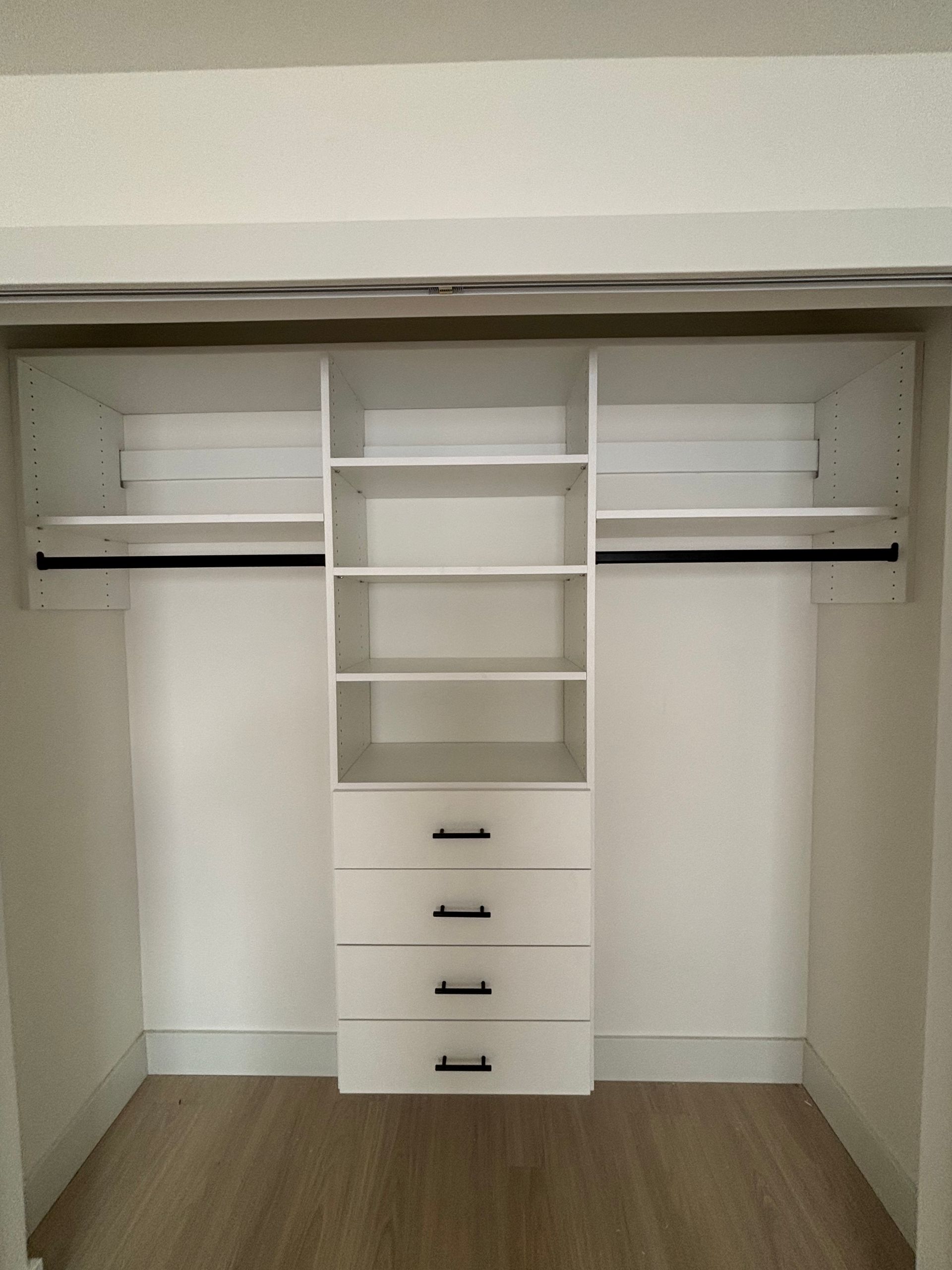 Empty walk-in closet with white shelving, hanging rods, and a central drawer unit on wood flooring