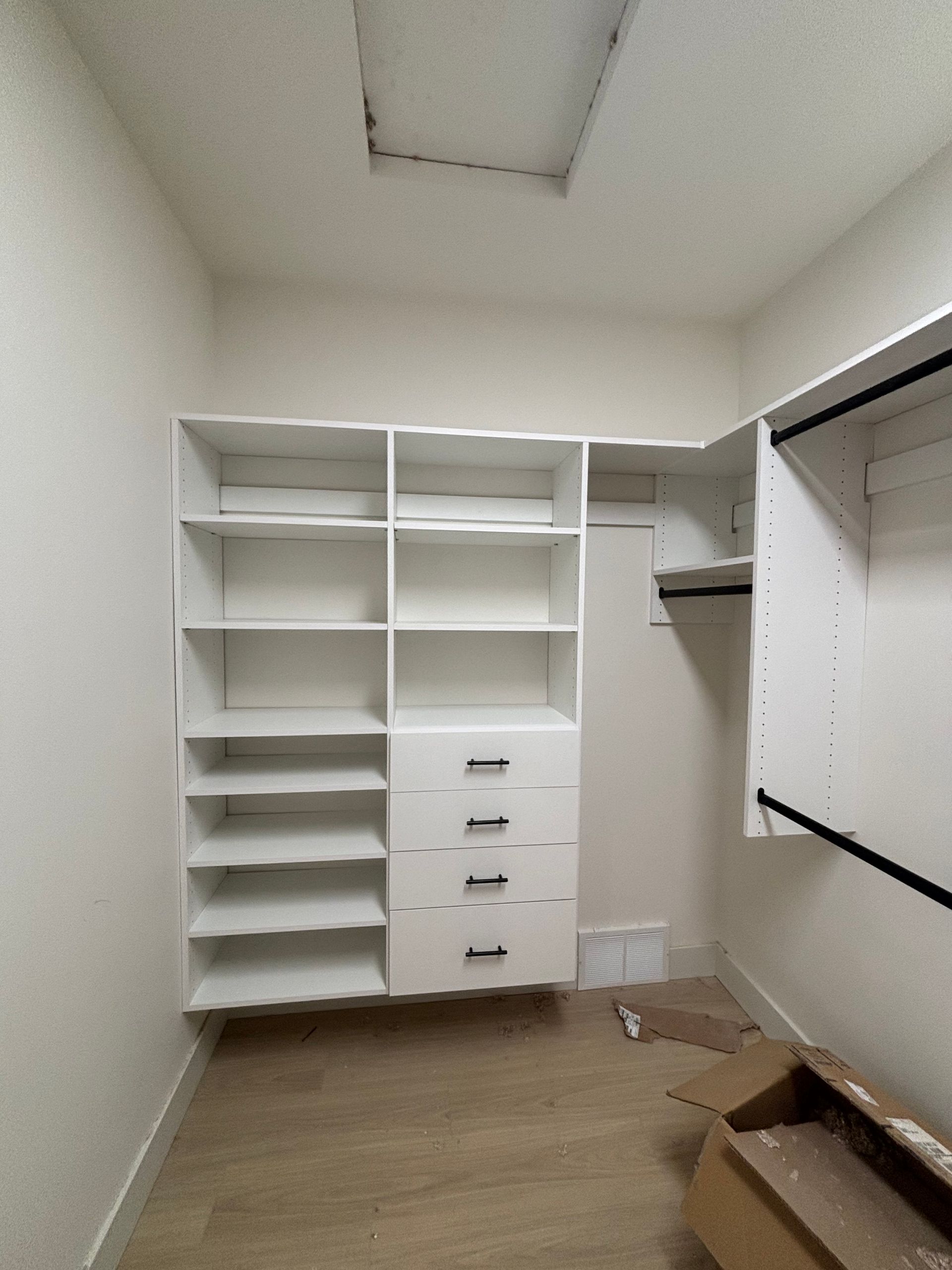 Empty white walk-in closet with shelves, drawers, and a hanging rod; cardboard box on the floor.