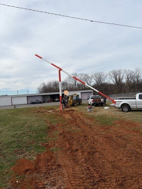 Barrier arm at a gate, red and white, blocking a dirt road. Two vehicles and equipment are nearby.