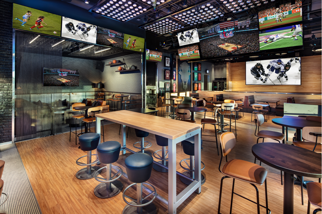 Sports bar interior with multiple screens showing games, tables, and bar stools.