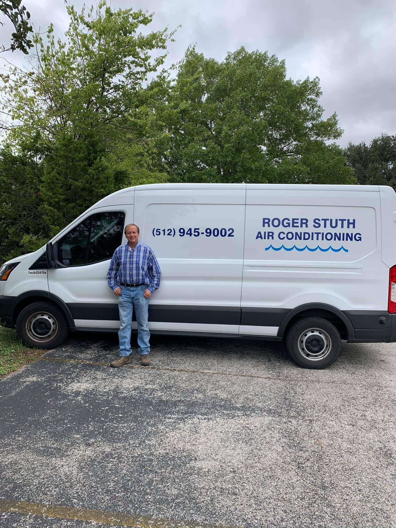 Man standing beside white Rodger Stuth Air Conditioning service van in a gravel lot with trees.