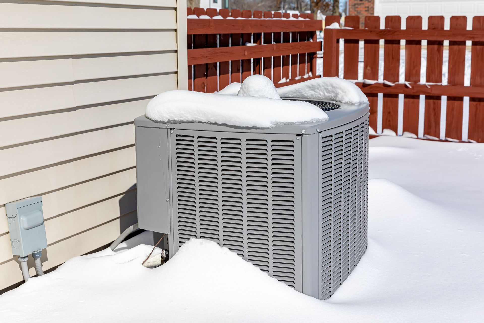 Gray air conditioning unit covered in snow next to a beige house, snow-covered yard, red fence.