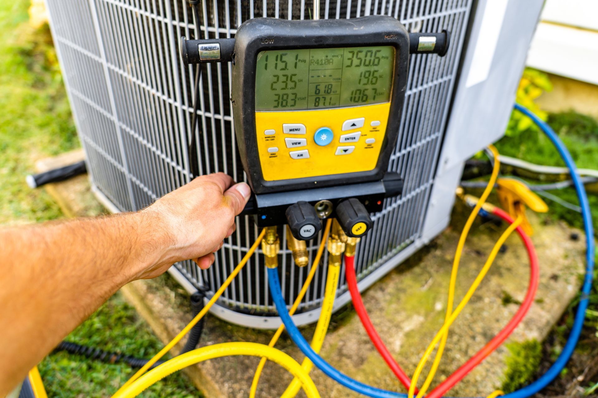 A technician's hand holds a digital manifold gauge connected to an outdoor HVAC unit with colorful hoses.