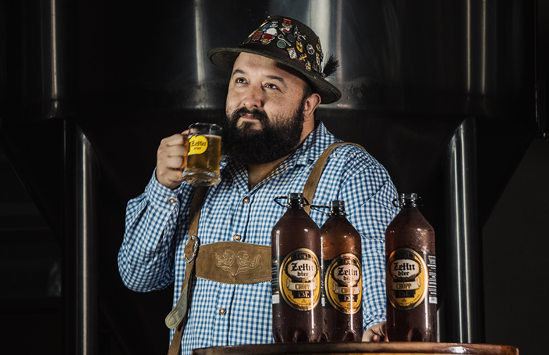 Um homem com barba segura um copo de cerveja na frente de três garrafas de cerveja.