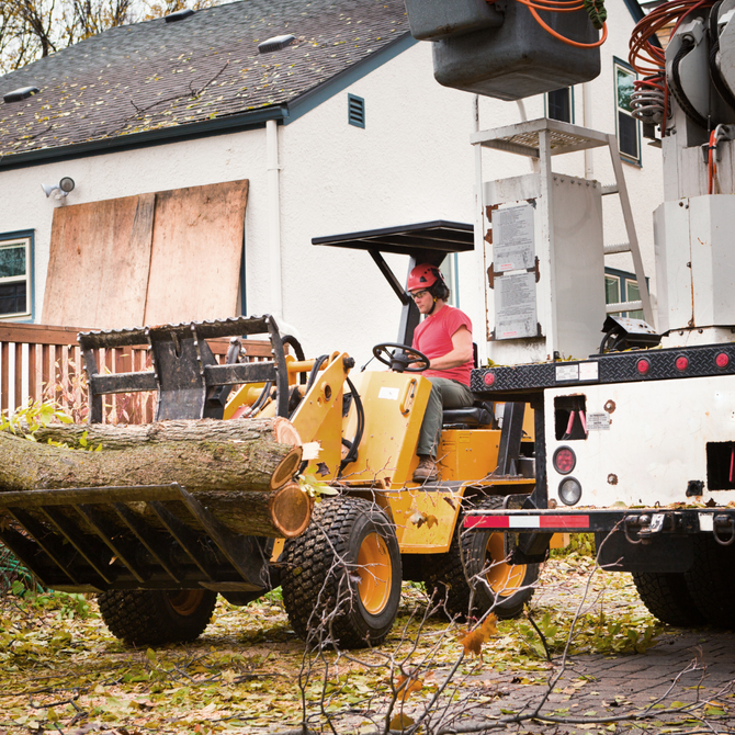 Yellow loader carrying logs at a worksite beside a truck and boarded-up building