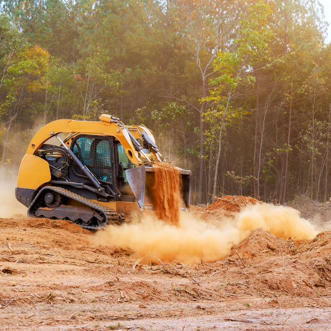 Yellow tracked skid steer dumping dirt in a muddy clearing with trees in the background