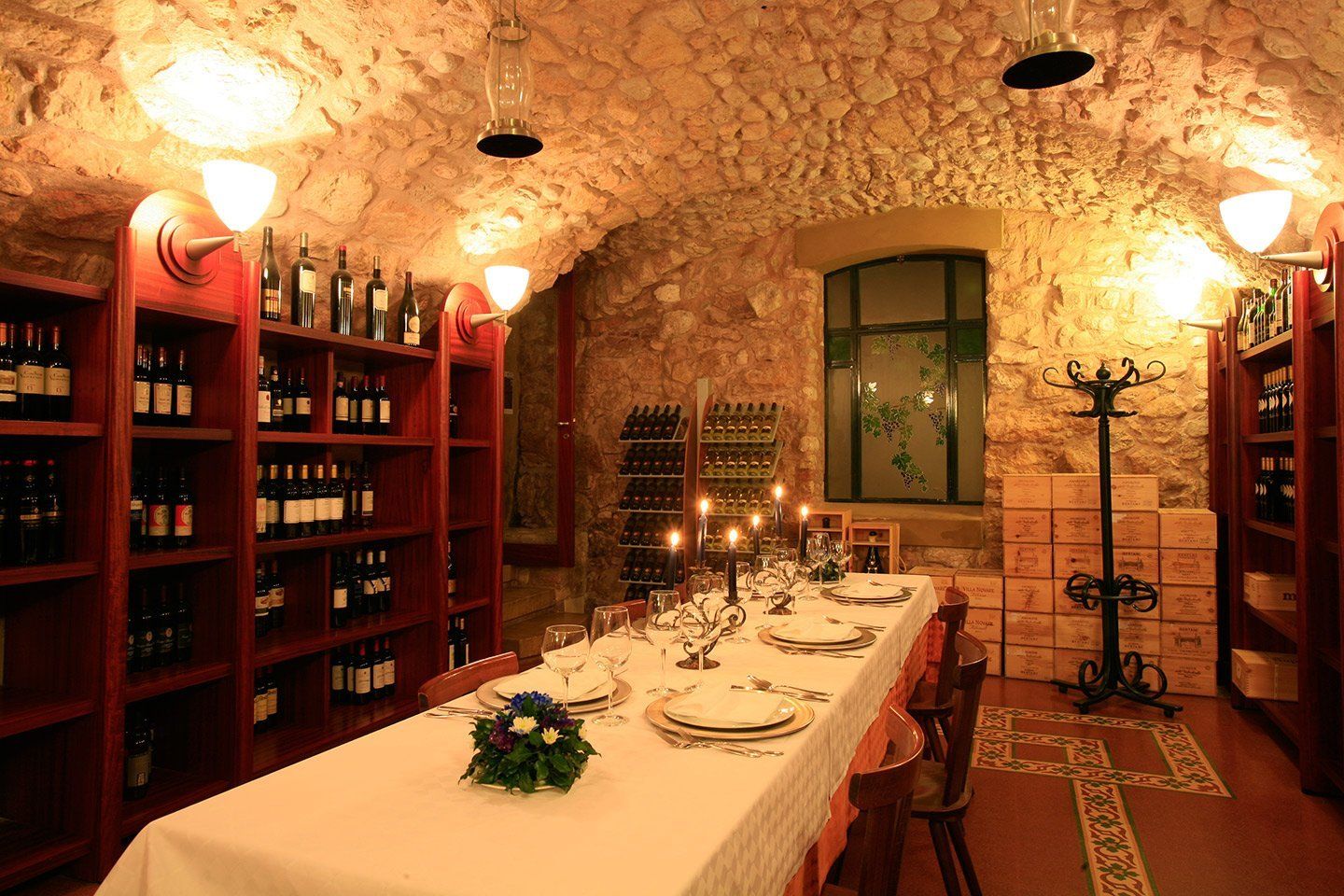 Sala da pranzo in cantina, illuminata da una luce calda, con un lungo tavolo apparecchiato, scaffali di bottiglie di vino e pareti in pietra.