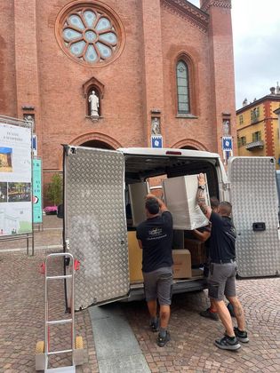 Due operai caricano un camion davanti a una chiesa in mattoni, con la strada bagnata e il cielo coperto.