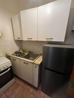 Piccola cucina con mobili bianchi, fornello a gas, lavello e frigorifero nero.
