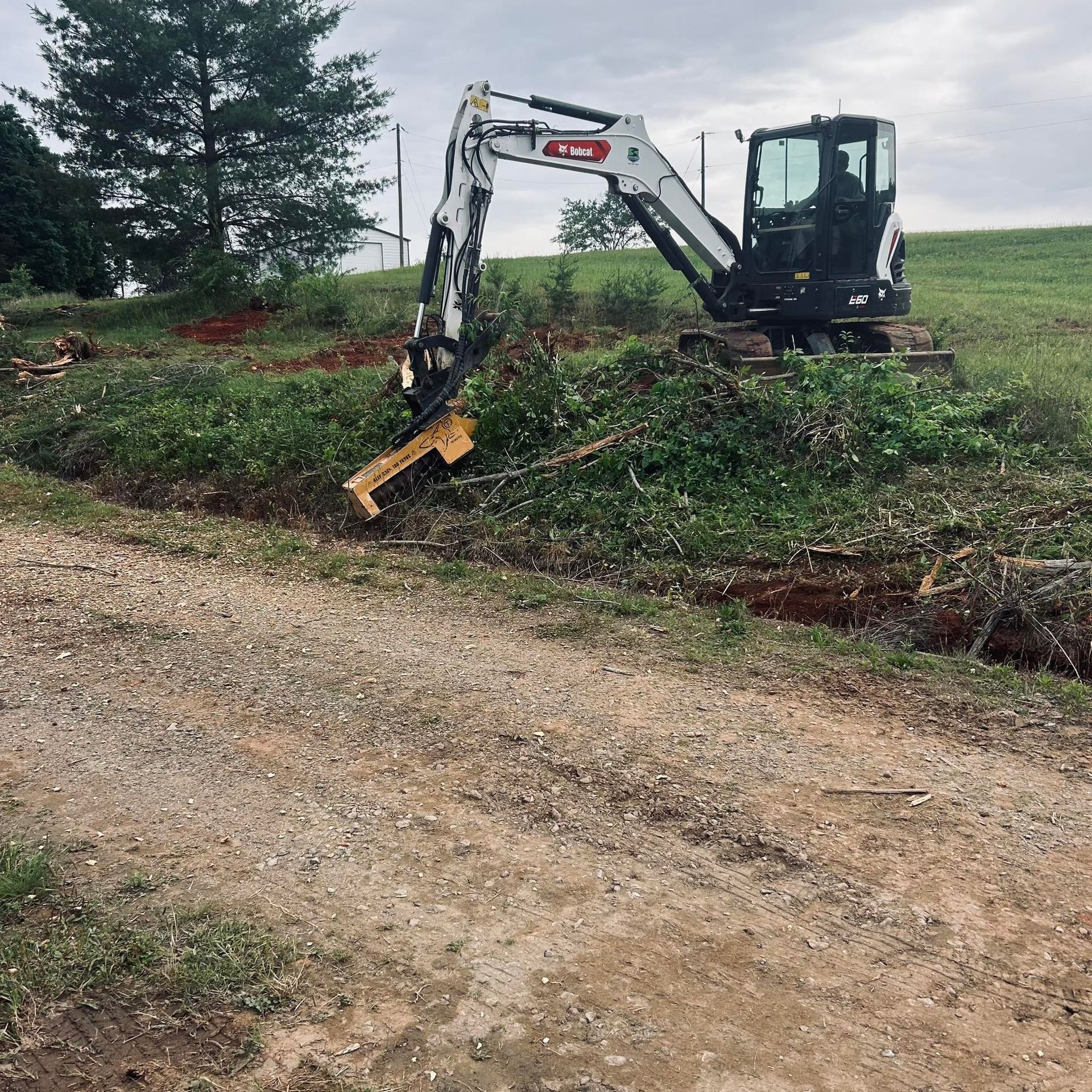 Excavator clearing brush beside a dirt road on a cloudy day