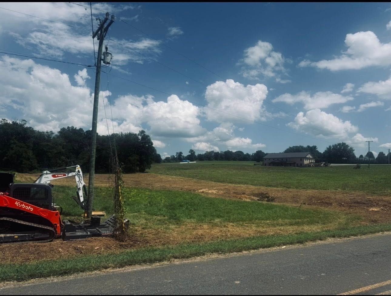 Rural roadside field under a cloudy sky, with a utility pole and excavator at the left edge