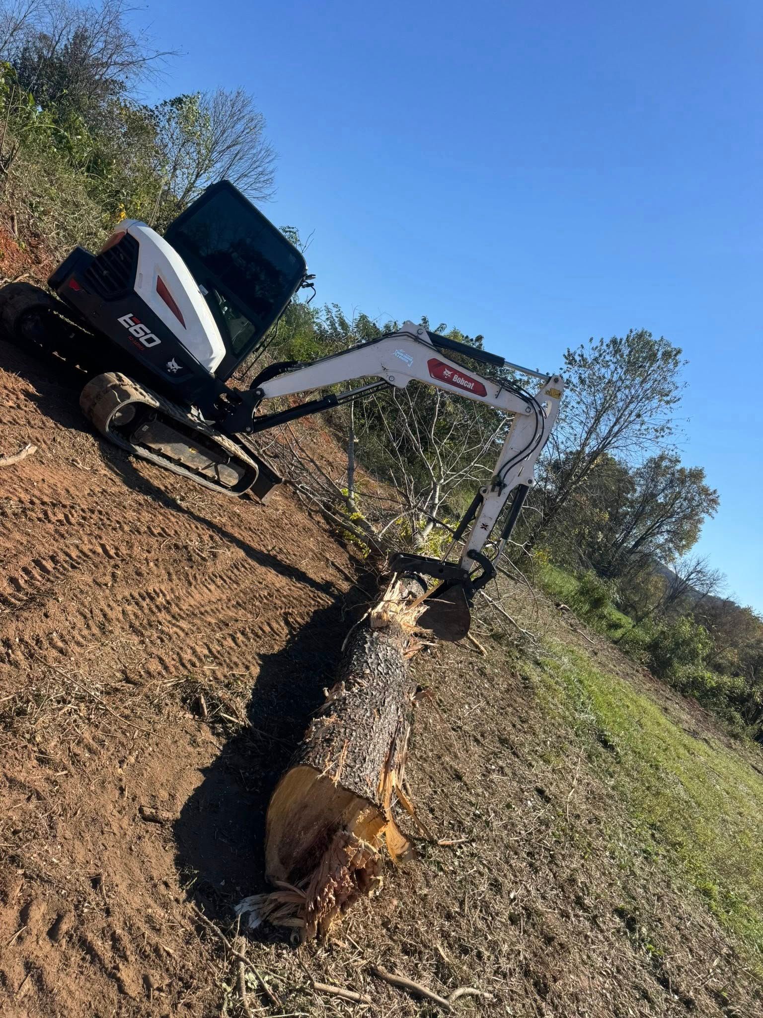 Excavator digging a trench in a dry, wooded field under a clear blue sky.