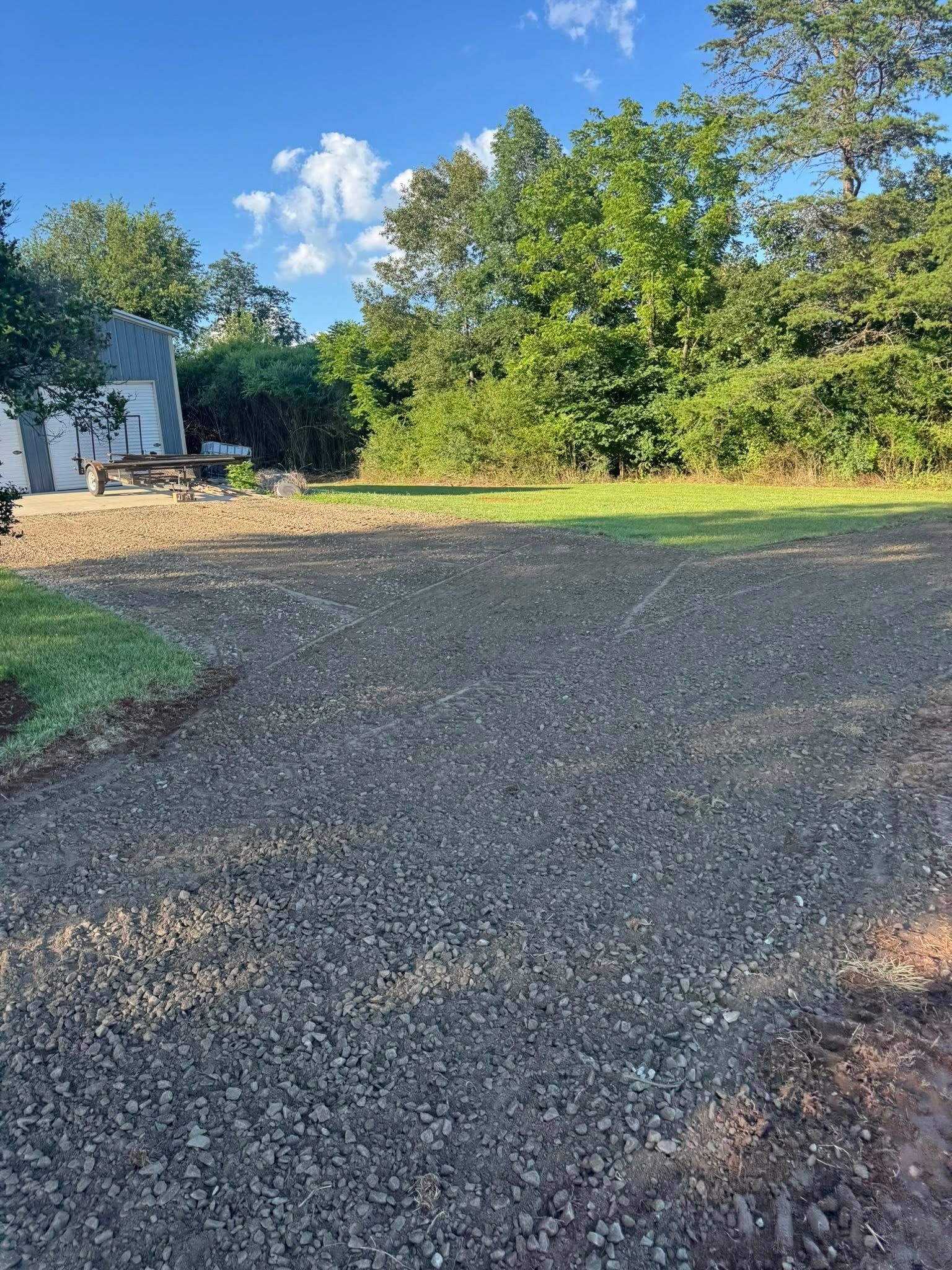 Gravel driveway beside green grass and trees under a bright blue sky with clouds