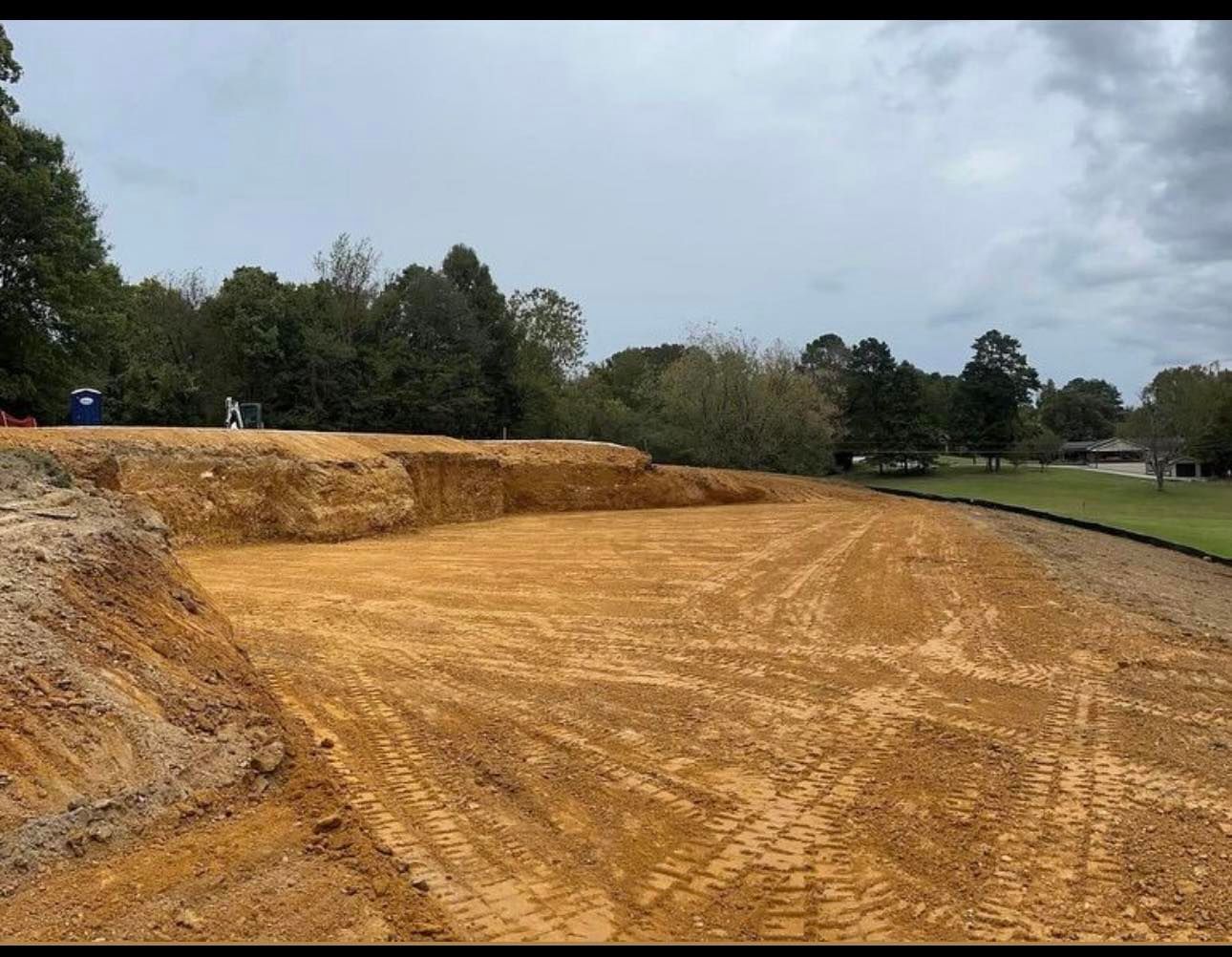 Freshly graded dirt construction site beside trees and a road under a cloudy sky