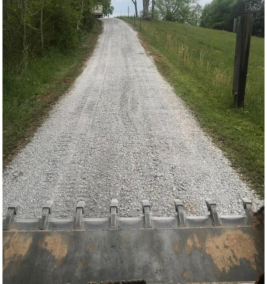 Gravel driveway rising uphill through a green field, with a metal equipment edge in the foreground