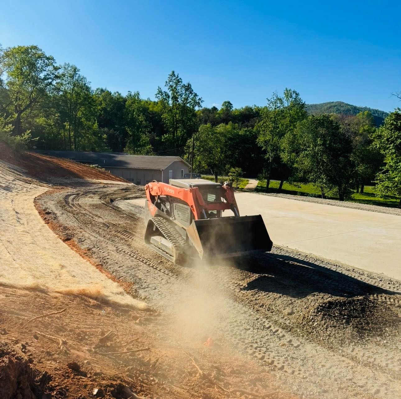 Orange skid-steer loader spreading gravel on a sunlit road construction site, kicking up dust