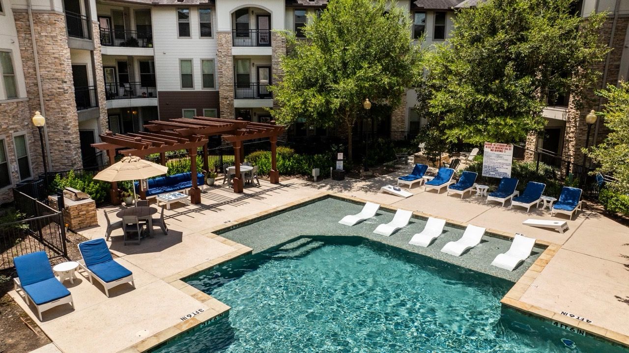 Outdoor apartment community pool with blue-cushioned lounge chairs and a wooden pergola at Olympus at Waterside Estates, offering luxury apartments in Richmond, TX.