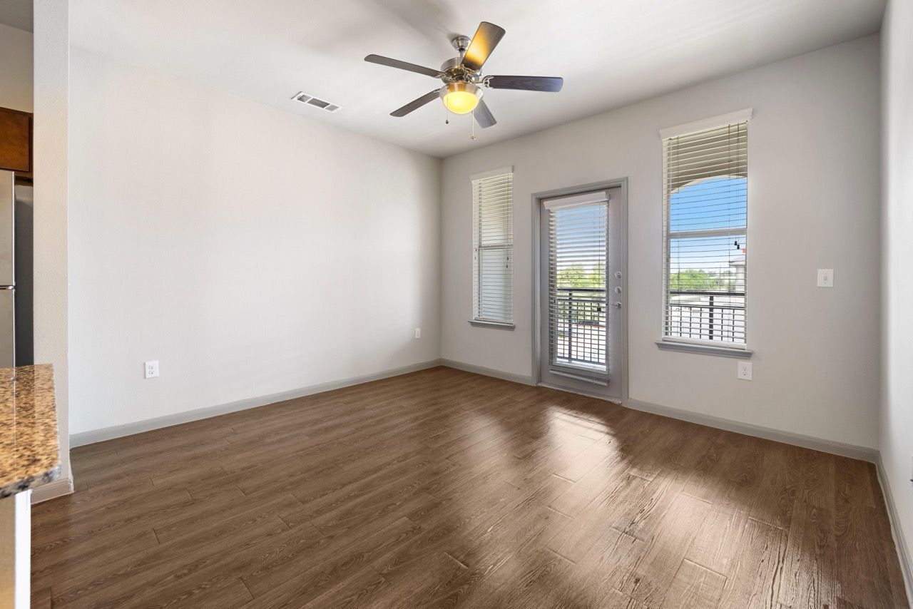 Empty living room with wood-look flooring, a ceiling fan, and a balcony door at Olympus at Waterside Estates, offering apartments in Richmond, TX.