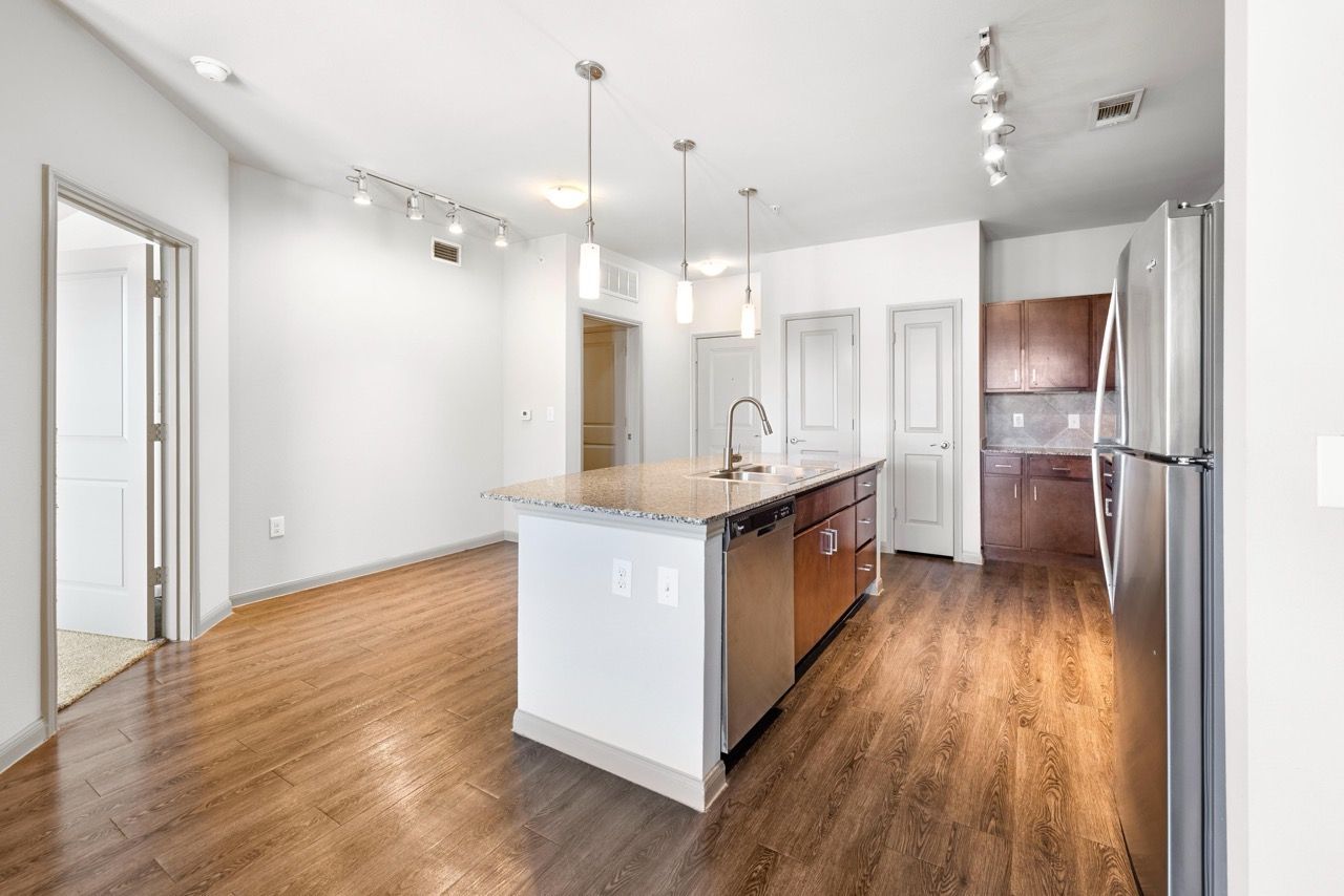 Open-concept kitchen with island, stainless fridge, pendant lights, and wood flooring at Olympus at Waterside Estates, offering apartments in Richmond, TX.