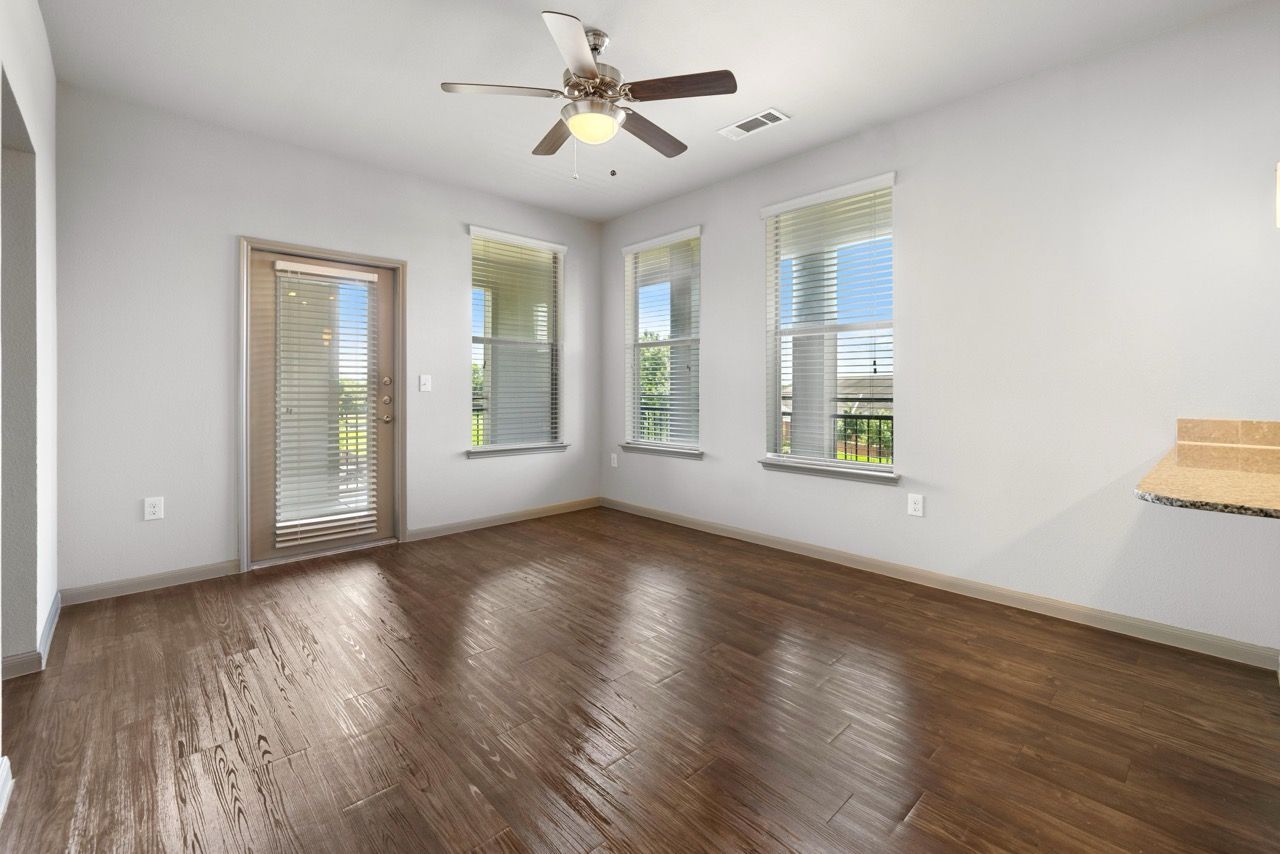 Living room with wood-look flooring, ceiling fan, and large windows with a balcony door at Olympus at Waterside Estates, offering apartments in Richmond, TX.